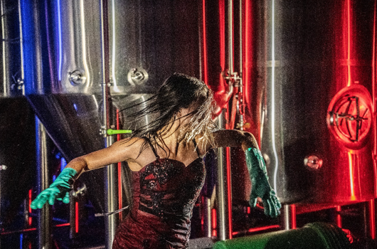 Une femme en robe, avec des gants de vaisselle, la face cachée derrière les cheveux, danse devant des cuves