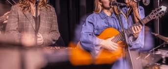 Deux chanteuse, une tenant une guitare classique, un percussionniste en arrière plan