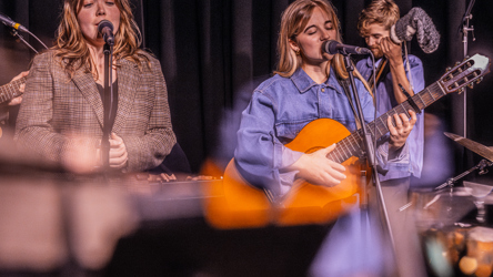 Deux chanteuse, une tenant une guitare classique, un percussionniste en arrière plan