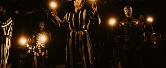 Des individus en costumes noirs et blancs, sur scène, qui tiennent des globes de lumière dans leurs mains