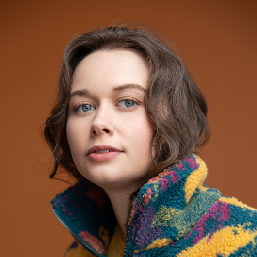 Femme aux yeux bleus et aux cheveux bruns, sur fond orangé, avec gilet multicolore