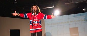 Un homme aux cheveux longs et vêtu d'un gilet de l'équipe de hockey des Canadiens de Montréal parle devant un micro, les bras ouverts