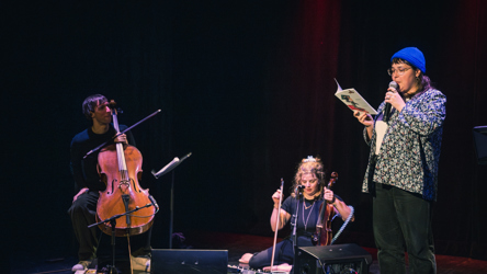 Une femme tient un micro et lis un livre, accompagnée de deux musiciens