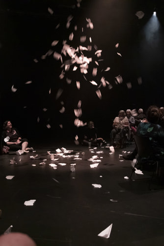 Un déversement de papier tombe sur un plancher sur lequel sont assis des individus en cercle